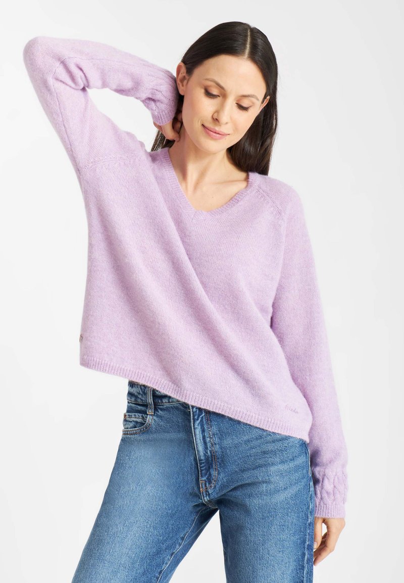 Vrouw in een lichtpaarse V-hals trui met kabelgebreide manchetten en blauwe jeans, poserend met één hand achter haar nek.