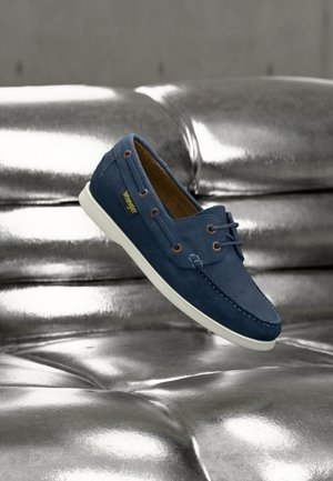 Mocasini albăstri din piele întoarsă cu talpă albă și găuri pentru șireturi maro, plutind deasupra unei canapele lucioase din piele argintie.