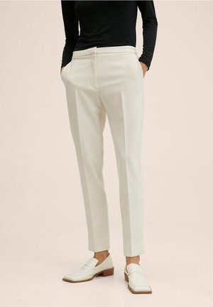 Personne portant un pantalon blanc sur mesure, des mocassins blancs et un haut noir à manches longues, debout les mains dans les poches.