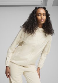 Ljust beige huva, vindjacka med elastiska cuffs och sidofickor. Slätt, glänsande tyg som har en liten logotyp på bröstet.