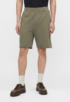 Mann trägt olivgrüne knielange Shorts, weiße Socken und dunkelbraune Leder-Schnürschuhe, steht vor einem einfarbigen Hintergrund.