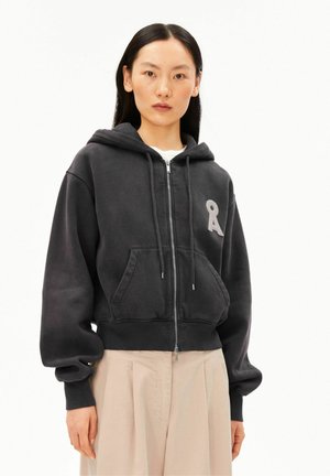Junge Frau mit langen dunklen Haaren, die einen schwarzen Kapuzenpullover mit kleinem Buchstaben-A-Emblem und beige weite Hosen trägt.