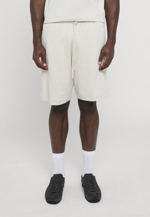Man wearing textured off-white shorts with drawstring, white crew socks, and black sneakers, standing against a plain light background.