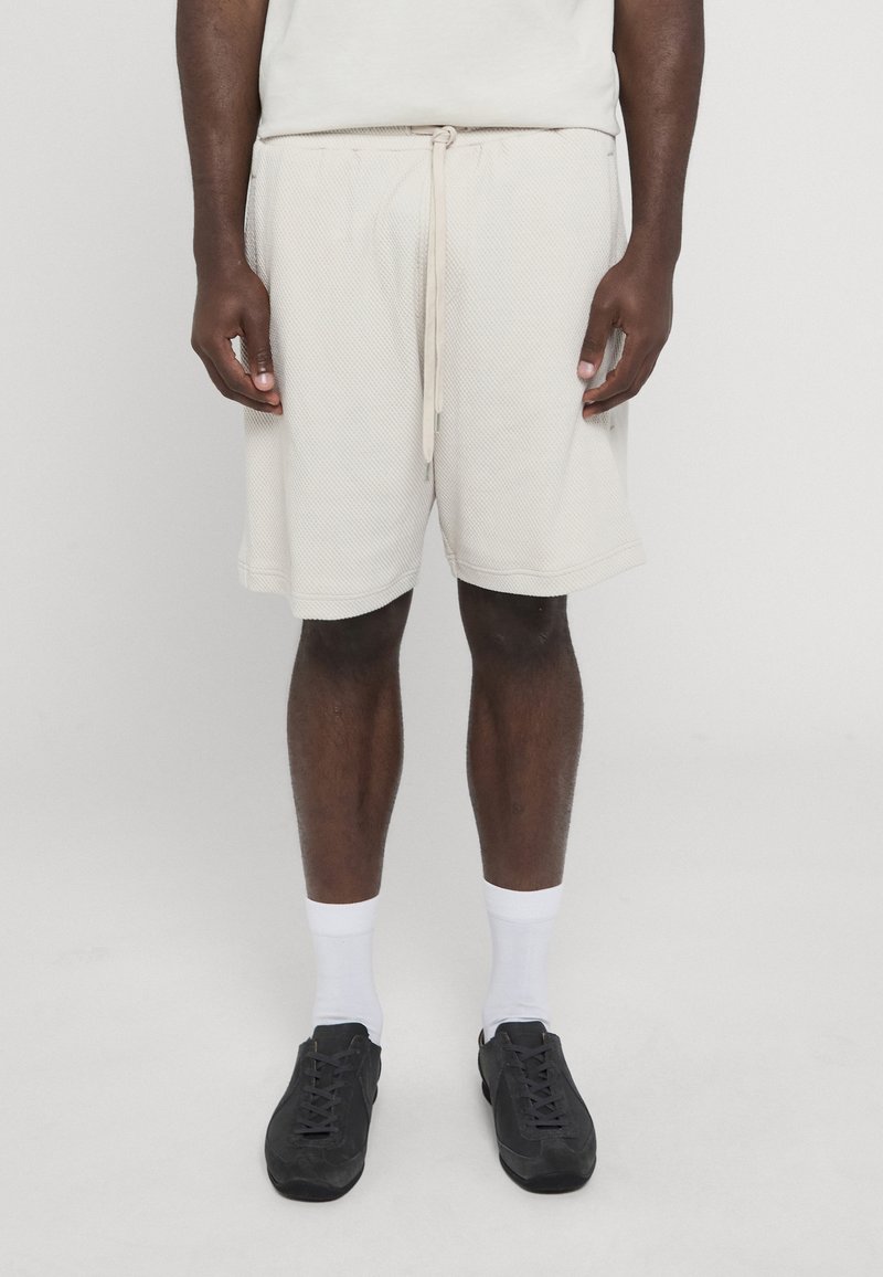 Homme portant un short écru texturé avec cordon, chaussettes blanches montantes et baskets noires, debout devant un fond clair uni.