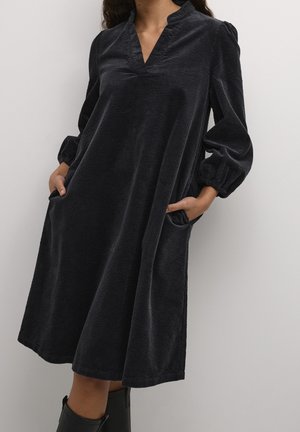 Femme portant une robe midi en velours côtelé noire avec de longues manches bouffantes, un décolleté en V et les mains dans les poches latérales, debout devant un fond uni.
