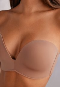 Tvarovaný podprsenkový bezramínkový model v jemné béžové barvě, vyznačující se hladkou texturou, bezšvovým designem a podpůrnými kosticemi s nízkým výstřihem.
