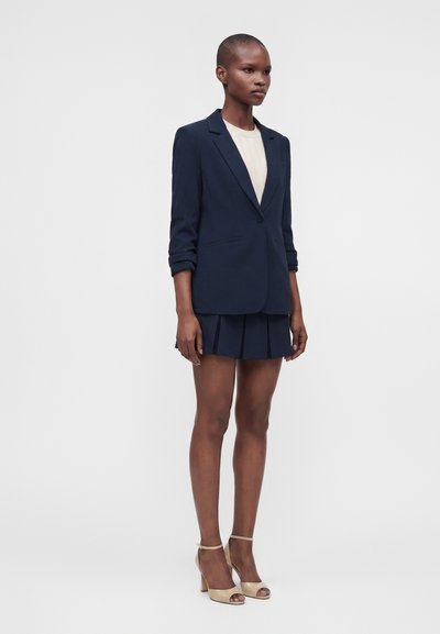Femme portant un blazer bleu marine avec les manches retroussées, un top blanc, une mini-jupe plissée bleue marine et des chaussures beiges à talons hauts ouvertes à l'avant, debout devant un fond blanc.