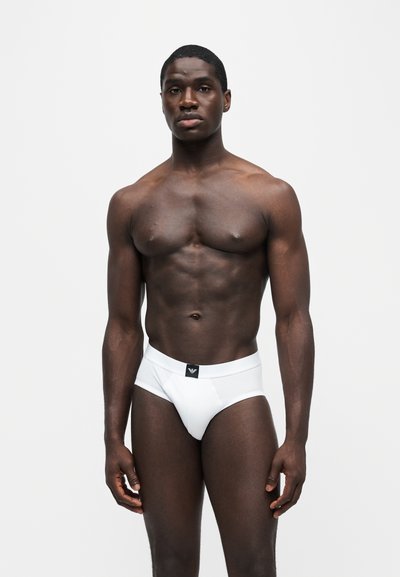 Homme musclé se tenant devant un fond uni, portant un slip blanc avec un logo noir sur la ceinture.