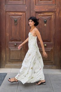 Biela maxi šaty s kvetinovým vzorom a ramienkami, voľná látka, zdobené nenápadnými zelenými a ružovými vzormi, v kombinácii s čiernymi sandálmi na podpätku.