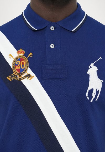 Kék piké ing három gombbal, fehér csíkkal és tengerészkék részletekkel. Fehér színű hímzett korona, "20" és pólójátékos logó díszíti.
