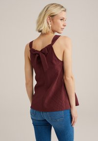 Burgundin väri tanktoppi, jossa on teksturoitu pinta, leveät hihnat ja sulkija selässä, yhdistettyinä kapeisiin sinisiin farkkuihin.