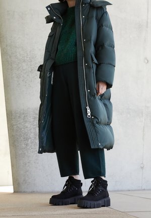 Abrigo acolchado largo verde con cuello alto y cierre de cremallera, combinado con un suéter de punto verde oscuro y pantalones negros cortos. Botas de plataforma negras.