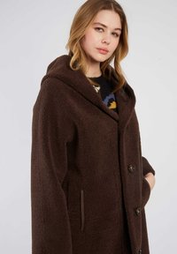 Brązowy płaszcz z polaru z kapturem, charakteryzujący się miękką fakturą, dużymi guzikami i głębokimi kieszeniami bocznymi. Zaprojektowany z myślą o ciepłu i komforcie.