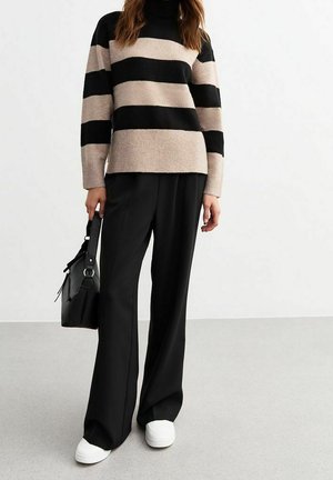 Personne portant un pull rayé noir et beige, un pantalon noir à jambes larges, des baskets blanches, et tenant un sac à main noir à côté d'elle.