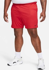 Uomo che indossa pantaloni sportivi rossi, calze bianche e scarpe Nike bianche, in piedi su uno sfondo bianco.