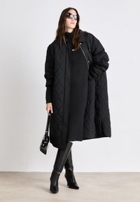 Svart vadderad lång kappa med avslappnad passform, bärs över en svart ribbad klänning, tillsammans med svarta knähöga spetsiga stövlar och en liten handväska.