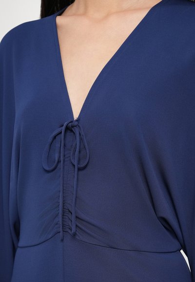 Ένδυμα σε ναυτικό μπλε με V-λαιμόκοψη και λεπτομέρεια δέσιμου, ρυτιδώσεις στο στήθος και λεία, ελαφριά υφή υφάσματος.