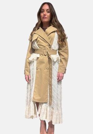 Donna con capelli castani lunghi che indossa un impermeabile beige con cintura sopra un vestito in pizzo bianco strutturato, in piedi davanti a uno sfondo chiaro e semplice.