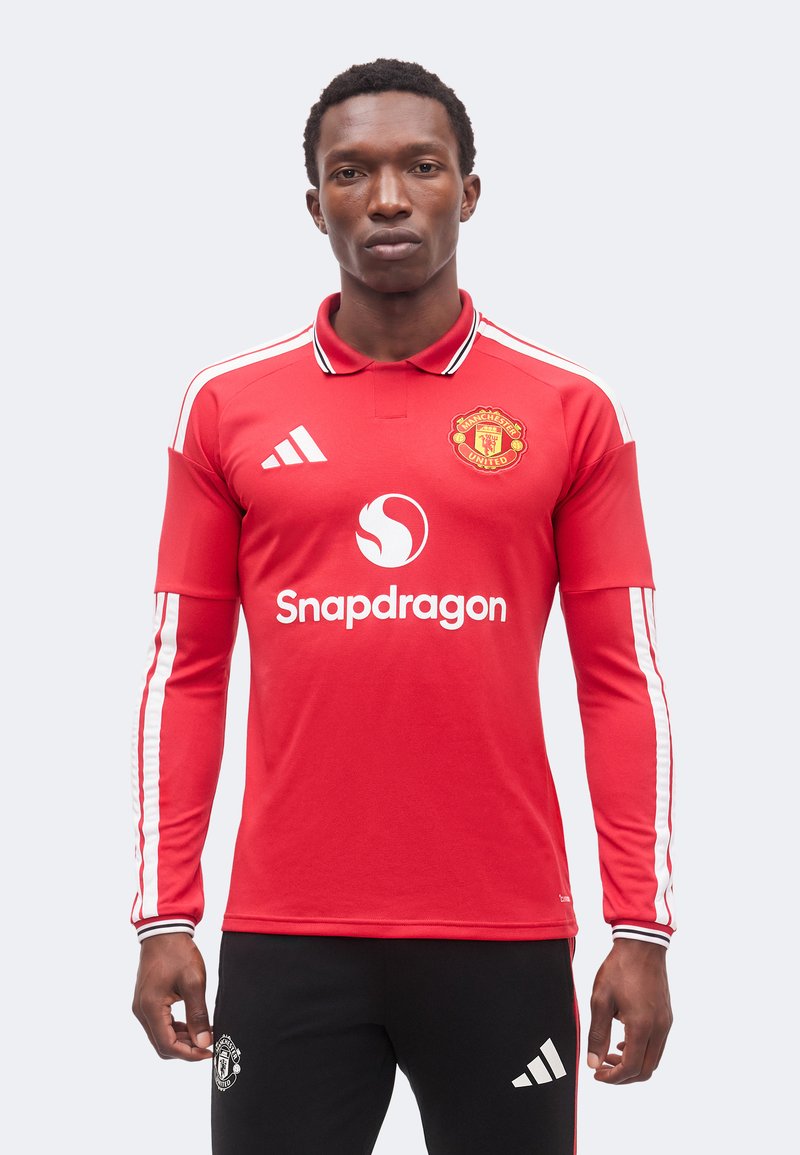 Άνδρας με κόκκινη φανέλα ποδοσφαίρου Manchester United με μακρύ μανίκι και λογότυπο "Snapdragon" και μαύρο παντελόνι Adidas σε απλό φόντο.
