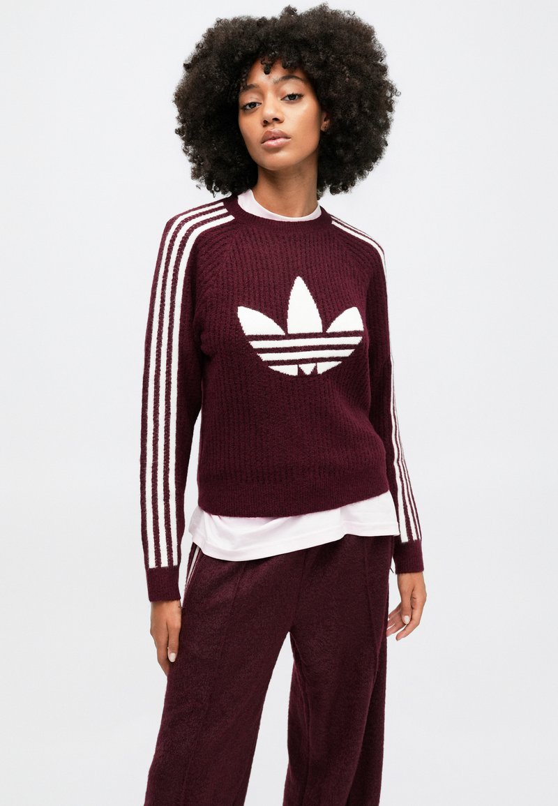 Burgunderfarbener Strickpullover mit weißen drei Streifen an den Ärmeln und einem großen weißen Trefoil-Logo auf der Vorderseite. Über einem weißen Hemd getragen.