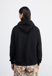 Black hoodie with a soft texture, featuring a prominent hood and ribbed cuffs, paired with patterned beige and brown pants.