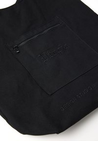 Bolso tote negro hecho de un tejido duradero con un bolsillo con cremallera, marca en relieve y textura suave. Presenta una forma redondeada.