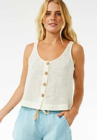 Weißes Leinen-Top mit Rundhalsausschnitt, Holzknöpfen entlang der Vorderseite und einem cropped Schnitt. Kombiniert mit hellblauen Shorts.
