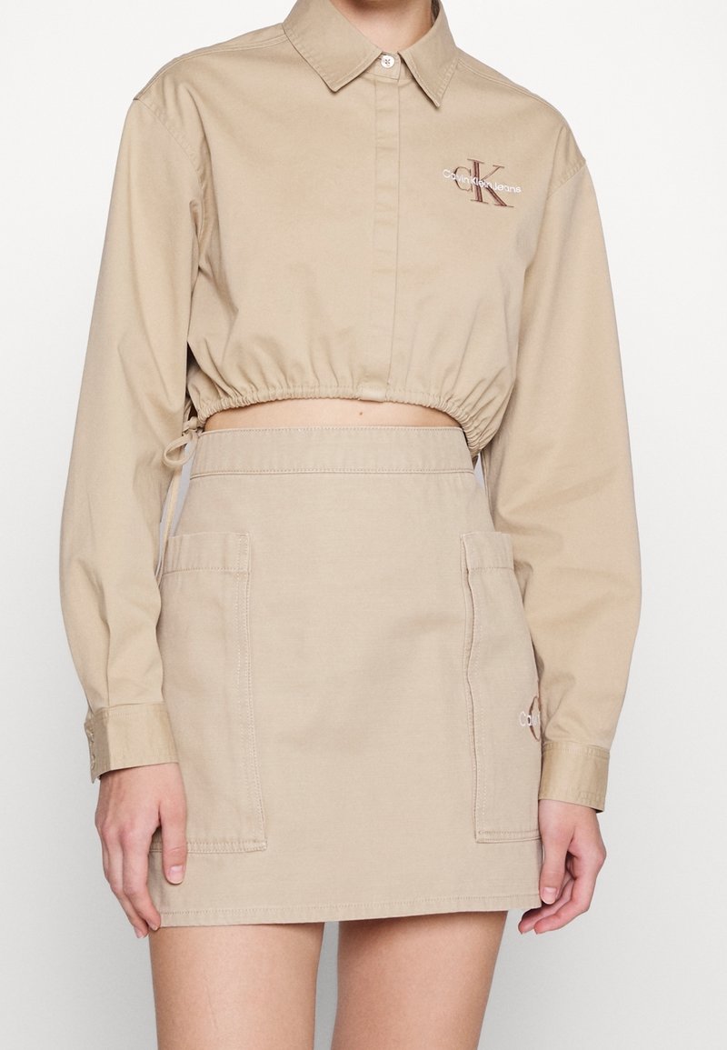 Chemise courte beige en coton, dotée d'une patte de boutonnage à l'avant et d'une ceinture élastique. Associée à une jupe beige assortie avec des poches.
