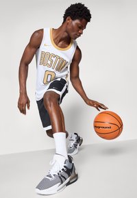 Witte basketbaltrui met gouden accenten, zwarte shorts en zwart/witte sneakers. Houdt een oranje basketbal vast, gefocuste houding.