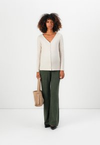 Kvinna med lockigt hår som bär en beige knappad cardigan, gröna byxor med bred ribb, svarta skor, håller en beige tote-väska, står på vit bakgrund.