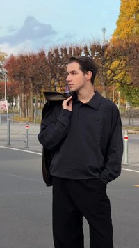 Camiseta polo negra de gran tamaño con un logo bordado sutil, combinada con pantalones negros sueltos y una chaqueta de cuero marrón oscuro sobre un hombro.