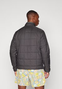 Veste matelassée noire avec col montant et couture visible, portée sur un short à motifs. Le matériau semble lisse et léger.