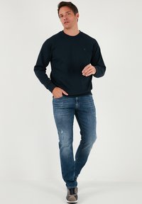 Tengerészkék pulóver kerek nyakkivágással és raglán ujjakkal, világoskék farmerrel, amely szakadt részletekkel rendelkezik, és szürke sportcipőkkel párosítva.