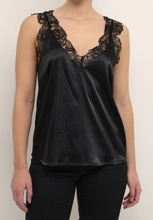 Kvinde iført sort satin camisole med blonder ved skuldre og halsudskæring, kombineret med sorte bukser, stående foran en ensfarvet baggrund.