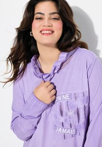 Paarse blouse met lange mouwen en gerimpelde halslijn, gaasaccenten en geborduurde tekstpatches. Heeft een relaxte pasvorm en een gestructureerde stof.