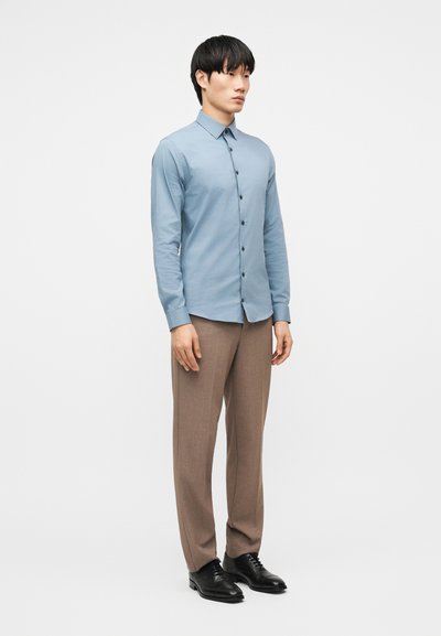 Άνδρας που στέκεται φορώντας ένα ανοιχτό μπλε πουκάμισο με κουμπιά, καφέ παντελόνια κ αι μαύρα επίσημα παπούτσια, μπροστά σε ένα απλό λευκό φόντο.