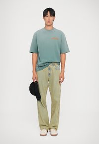 Camiseta de algodón verde con logo naranja, pantalones de mezclilla a rayas claras, zapatos blancos y beige, sosteniendo una gorra negra. Fondo liso.