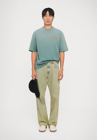 Groene katoenen T-shirt met oranje logo, licht gestreepte denim broek, witte en beige schoenen, houdt een zwarte pet vast. Effen achtergrond.