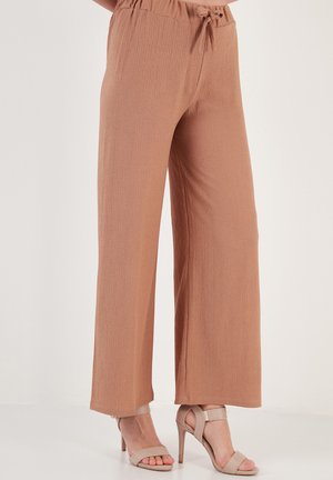Hellbraune, weit geschnittene Hose mit elastischem Bund und Kordelzug-Detail; strukturierter Stoff und glatte Oberfläche. Kombiniert mit beigen High-Heels-Sandalen.