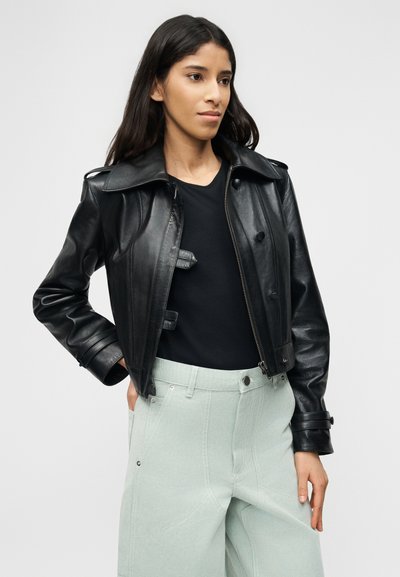 Γυναίκα που φορά ει μαύρο δερμάτινο μπουφάν πάνω από μαύρο τοπ και ανοιχτό πράσινο-μίντ ψηλόμεσο παντελόνι, στέκεται με το ένα χέρι στην τσέπη.