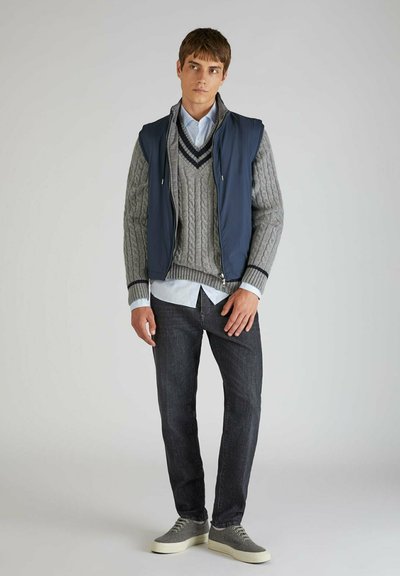 Chaleco sin mangas azul marino con cremallera sobre un suéter gris de punto trenzado con escote en V, superpuesto a una camisa de botones azul claro y jeans oscuros. Zapatillas grises.