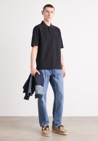 Sort polo-shirt med korte ærmer og broderet logo, parret med lyseblå denimjeans og beige sko, mens han holder en denimjakke.