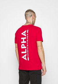 Crvena pamučna majica s velikim bijelim natpisom "ALPHA INDUSTRIES" vertikalno na leđima. Kratki rukavi, okrugli izrez i opušteni kroj.