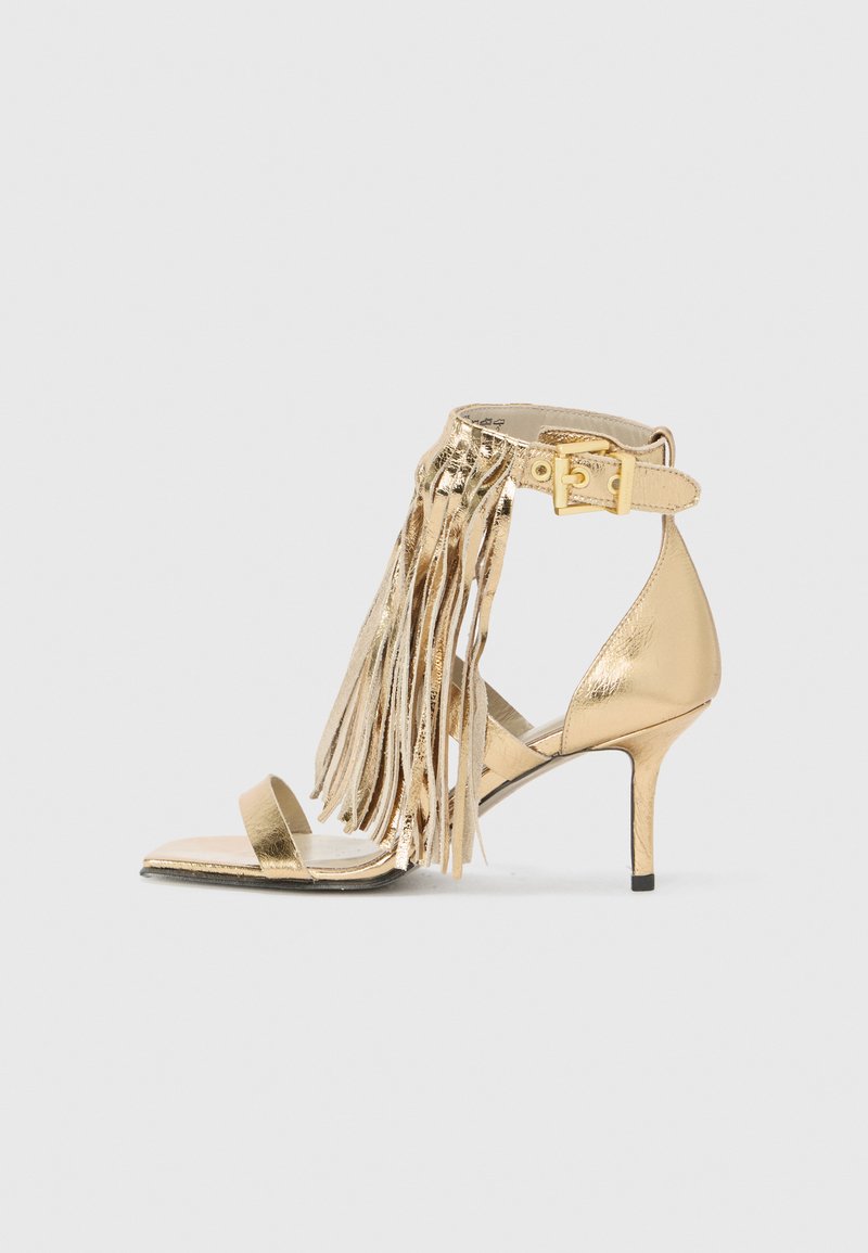 Gold high-heeled sandal featuring fringe detailing, an ankle strap with a buckle, and a slim stiletto heel. Smooth metallic finish.