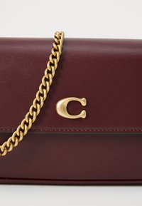 Burgunderfarbene Ledertasche mit glatter Textur, ausgestattet mit einem goldenen Kettenriemen und einem auffälligen goldenen "C"-Logo auf der Vorderseite.