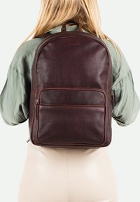 Sac à dos en cuir marron avec finition texturée, forme rectangulaire, poche zippée à l'avant et sangles réglables. Porté au dos, avec un minimum de décorations.