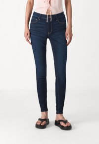 Mörkblå skinny jeans med en slät textur, medelhög midja och femficksdesign. I kombination med en ljusrosa topp och svarta sandaler.