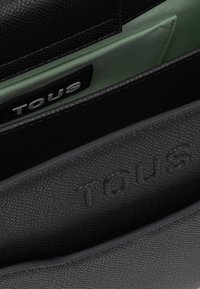 Zbliżenie na czarną torebkę skórzaną o fakturze z wytłoczonym napisem "TOUS" na zewnątrz oraz z tkaninową metką z napisem "TOUS" wewnątrz.