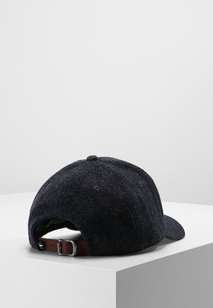 Casquette de baseball en laine gris foncé avec une sangle réglable en cuir marron et une boucle en métal, posée sur une surface blanche devant un fond gris clair.