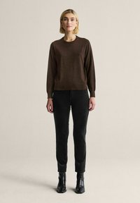 Ruskea neule pitkähihaisilla hihoilla, pyöreällä pääntiellä ja pehmeällä tekstuurilla, yhdistettynä mustiin slim fit -farkkuihin ja mustiin nilkkureihin. Neutraali tausta.